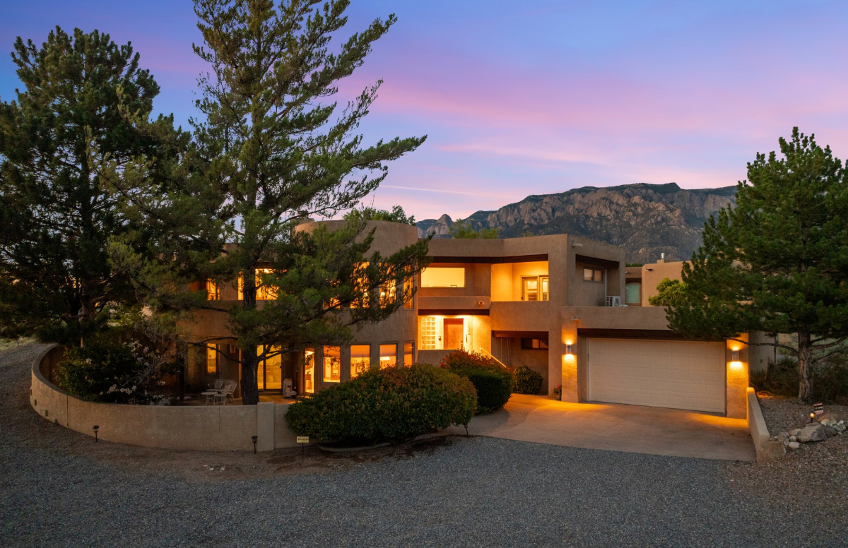 Two-story home at twilight in the Foothills of ABQ, NM, with mature pine trees and the Sandia Mountains behind it.