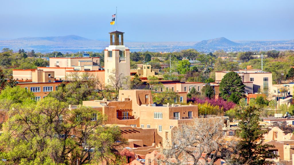 Aerial view of downtown Santa Fe, New Mexico, showing adobe buildings around the plaza with distant mountains beyond.