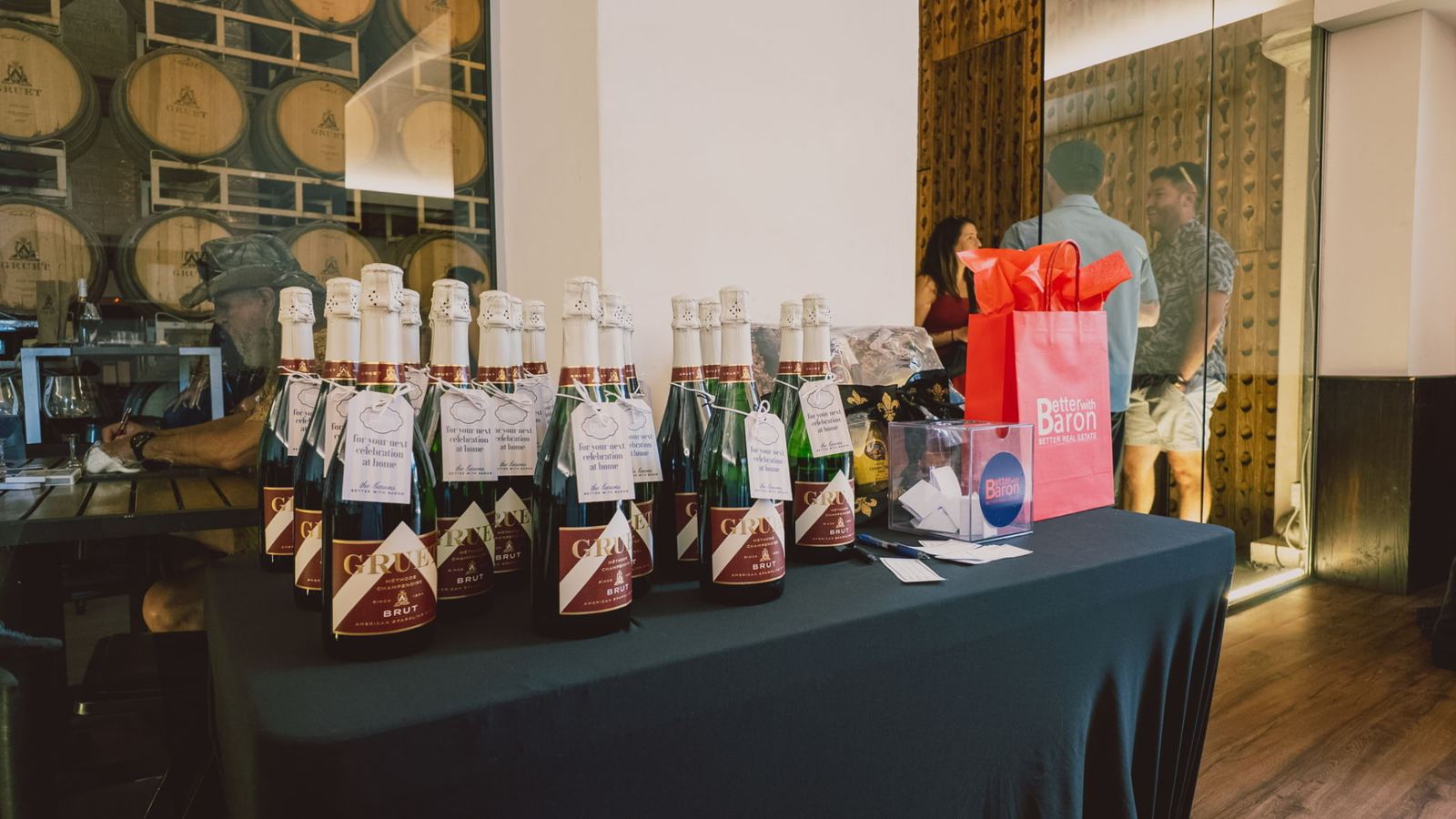 Gruet brut sparkling wine bottles with Better with Baron gift tags and a branded gift bag at an Albuquerque client appreciation party.