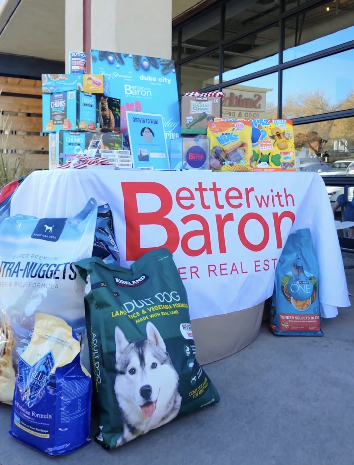 Better with Baron branded table stacked with donated dog food and pet supplies at a holiday pet food drive in Albuquerque.