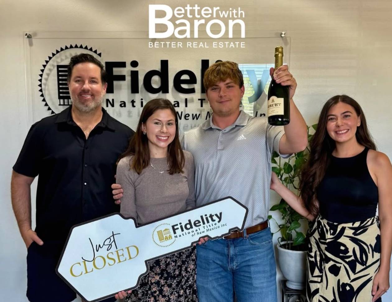 Two happy clients holding a large key that reads "just closed" and a bottle of champagne. They are with the Better with Baron Team who just helped them buy a home. buying a house in Albuquerque this summer