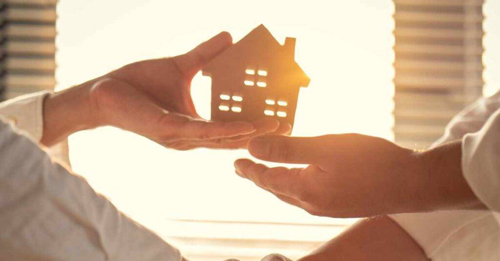 Two hands shaking and the other two exchanging a small wooden house. Seller financing Albuquerque.